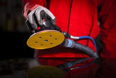 Uma pessoa usando luvas de segurança cinza segura uma lixadeira orbital pneumática equipada com um disco de lixamento amarelo. A parte inferior do disco está visível, mostrando múltiplos furos para extração de pó dispostos ao redor do centro. O corpo preto e vermelho da lixadeira, incluindo um logotipo “3M” visível, está conectado a uma mangueira de ar azul. A ferramenta está posicionada acima de uma superfície preta refletiva que espelha o disco e a mangueira.