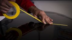 Close-up de uma mão aplicando fita de mascaramento amarela a partir de um rolo sobre uma superfície preta refletiva, guiando a fita diagonalmente enquanto a pressiona para formar uma linha reta e limpa.