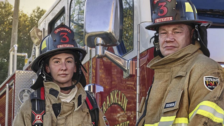 Two firefighters in turnout gear standing in front of a rescue fire truck, wearing helmets and SCBA equipment.