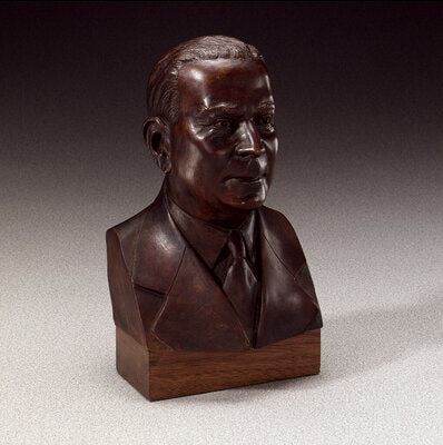 Small bronze bust of a suited figure on a rectangular base, displayed against a neutral studio background.