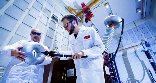 Two individuals wearing white lab coats with the 3M logo are working with electrical equipment in a laboratory setting.